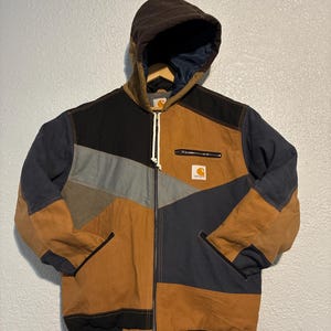 May include: A patchwork jacket with a brown hood and a full-length zipper. The jacket features a color-block design with brown, black, gray, and navy blue panels. A small Carhartt logo is visible on the chest.