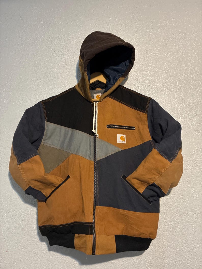 May include: A patchwork jacket with a brown hood and a full-length zipper. The jacket features a color-block design with brown, black, gray, and navy blue panels. A small Carhartt logo is visible on the chest.