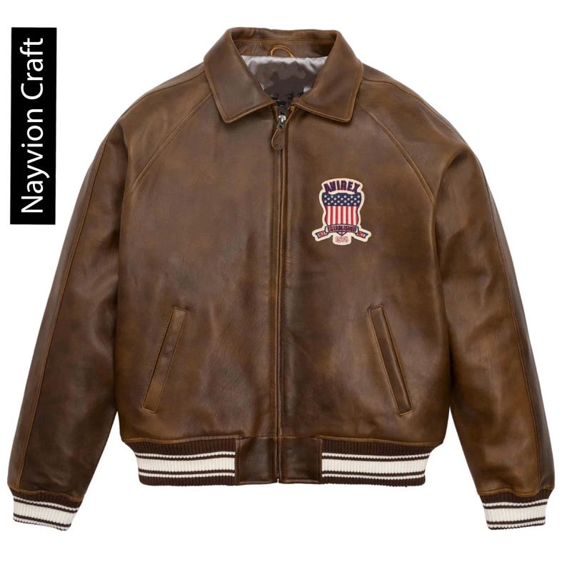 May include: Brown leather jacket with a collared neckline, zipper closure, and a patch on the chest. The jacket features a ribbed hem and cuffs with white stripes. The text "Nayvion Craft" is on the left side.