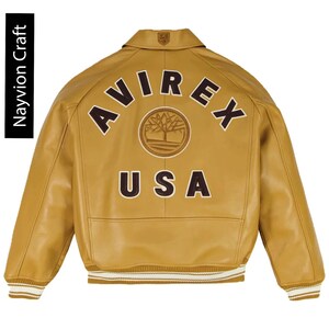 May include: A tan leather jacket with the word "AVIREX" arched across the back in brown letters, with "USA" below. The jacket has a ribbed collar and cuffs with white stripes. The brand name "Navyion Craft" is printed vertically on the left side.