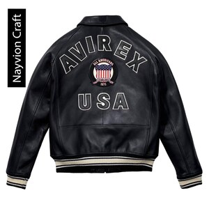 May include: Black leather jacket with white embroidered lettering and a patch featuring the American flag. The jacket has a classic bomber style with a ribbed hem and cuffs. The text "AVIREX" and "USA" are prominently displayed.