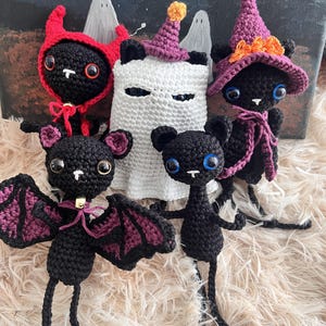 Puede incluir: Una colección de cinco figuras de ganchillo que incluye tres gatos negros, un fantasma blanco y un murciélago negro. Los gatos llevan una capucha de diablo roja, un sombrero de bruja morado y uno es sencillo. El murciélago tiene alas moradas.