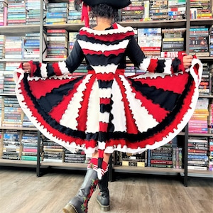 May include: A woman wearing a red, white, and black dress with a ruffled skirt and a black hat. The dress is made of knit fabric and has a fitted bodice. The woman is standing in front of a bookshelf.