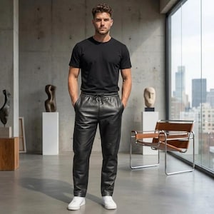 Könnte beinhalten: Eine Person trägt ein schwarzes Rundhals-T-Shirt, eine schwarze Lederhose mit Kordelzug und weiße Turnschuhe. Die Person steht in einem modernen Raum mit Skulpturen und Blick auf die Stadt.