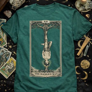 May include: Teal green t-shirt with a tarot card illustration. The design shows a person suspended upside down from a tree, with the words "THE HANGED MAN" beneath. The card is framed with a decorative border and the Roman numeral XII.