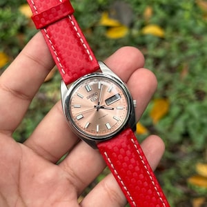 May include: A Seiko 5 automatic wristwatch featuring a round silver-tone stainless steel case and a metallic salmon-colored dial with silver hour markers. The watch is attached to a bright red textured strap with white contrast stitching.