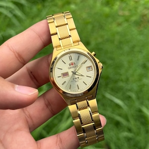 May include: A gold-tone Orient wristwatch with a textured champagne dial, featuring a day-date window at the three o'clock position, three-star logo, and a metal link bracelet with a central textured pattern on each link.