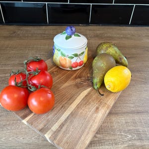 May include: A wooden chopping board displays a cluster of red tomatoes, two green pears, and a yellow lemon. A small, white ceramic jar with a fruit design and a blue berry on top is also present. The items are arranged on a wooden surface.