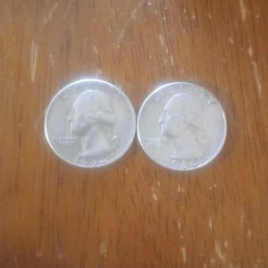 May include: Two silver 1964 quarter coins, each showing a profile of a person on the obverse. The coins are on a wooden surface. The coins are a United States currency.