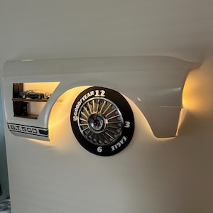 Puede incluir: Reloj de pared blanco con forma de guardabarros de coche, con una esfera de reloj de neumático negro. El reloj presenta las palabras "GOOD YEAR" y "EAGLE". Una pequeña estantería muestra modelos de coches, y el texto "GT.500" es visible.