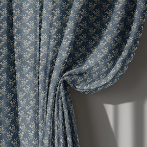 May include: A blue curtain panel featuring a repeating pattern of small, light-colored sprigs or leaves. The fabric has a visible weave texture and is gathered to one side, creating soft folds that drape vertically from the top to the bottom.