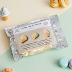 Op de afbeelding: Een pakket Gourmet Clay - Marshmallow, met miniatuur klei desserts, waaronder een stuk cake, een macaron en een ijsje. Twee macarons en een klein gebakje zijn ook zichtbaar.