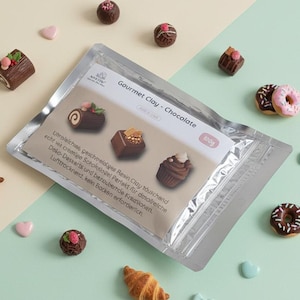 Op de afbeelding: Een pakket "Gourmet Clay - Chocolate" met miniatuur klei desserts, waaronder een croissant, donuts en chocolade snoepjes. Het pakket is zilver en wit, met tekst en afbeeldingen van de klei desserts. De achtergrond is lichtgroen en beige.