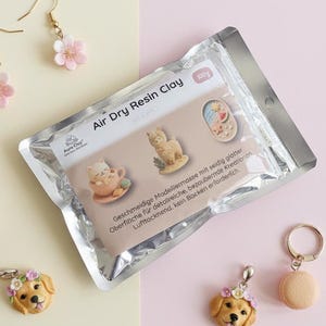 Op de afbeelding: Een assortiment handgemaakte polymeerklei sieraden en bedels, waaronder oorbellen met roze bloemhangers, een aardbeien ijsje bedel, een golden retriever bedel en een macaron sleutelhanger. Een verpakking luchtdrogende resin klei is ook aanwezig.