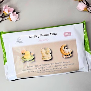 Könnte beinhalten: Eine 150g Packung Aura Clay Air Dry Foam Clay. Die weiße Verpackung zeigt auf einem beigen Mittelfeld drei kleine Tonfiguren: eine gelbe Ente, eine Schnecke und einen Hasen auf einem Mond. Der Text lautet Air Dry Foam Clay.