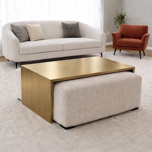 Könnte beinhalten: Ein modernes Wohnzimmer mit einem beige gepolsterten Ottoman und einem goldfarbenen rechteckigen Couchtisch. Ein passendes beiges Sofa mit Kissen und ein orangefarbener Sessel vervollständigen die Raumausstattung.
