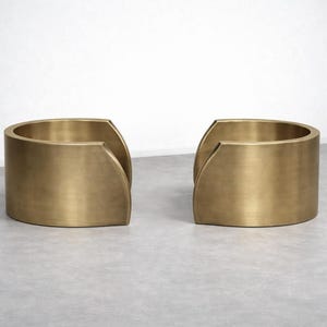 Op de afbeelding: Twee bijpassende, goudkleurige, halfronde bijzettafels. Elke tafel heeft een gebogen, cilindrisch lichaam met een uitgesneden gedeelte aan één kant. De tafels zijn gemaakt van massief metaal en staan op een neutrale ondergrond.