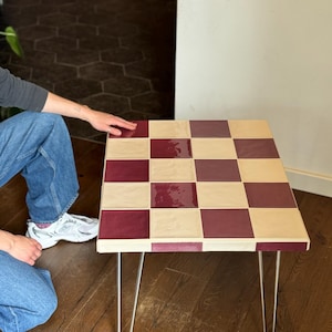 May include: A square side table with a checkerboard pattern in burgundy and cream tiles. The table has a light wood frame and thin, silver hairpin legs. The table is approximately 24 inches square.