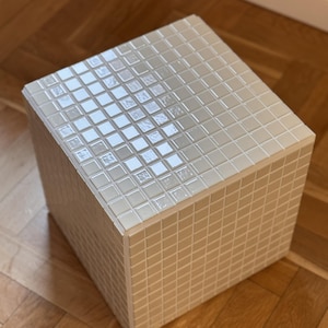 Może przedstawiać: Kwadratowy stolik boczny pokryty beżowymi kafelkami mozaikowymi. Górna powierzchnia ma gradient jaśniejszych płytek. Stół wykonany jest z małych, kwadratowych płytek, co nadaje mu teksturowany wygląd. Stoi na drewnianej podłodze. Wymiary około 40 cm x 40 cm x 40 cm.