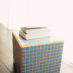 May include: A cube-shaped side table with a blue tile pattern and orange trim. Two white books are stacked on top. The table is on a wooden floor.