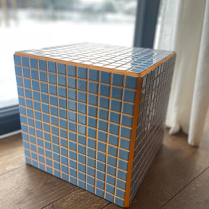 May include: A square side table with a mosaic tile design. The tiles are a muted blue color, set against a bright orange frame. The table is a cube shape and sits on a wooden floor.