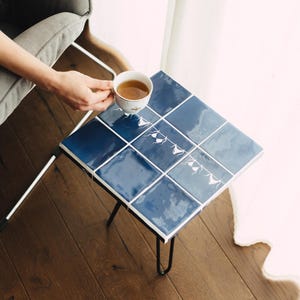 Peut inclure: Une table d'appoint carrée dont le plateau est composé de neuf carreaux de céramique bleu foncé brillants. Les carreaux présentent un dessin au trait blanc représentant du linge suspendu à une corde. La table repose sur quatre pieds en épingle.