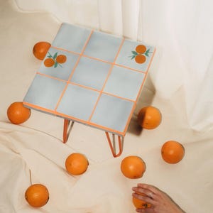 May include: A small, square table with a light blue tiled top and orange trim. The table has orange hairpin legs and is decorated with orange fruit illustrations. Oranges are scattered around the table.
