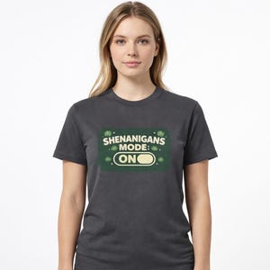 May include: Dark gray t-shirt with a green rectangle graphic. The graphic reads "SHENANIGANS MODE: ON" in white and green text. The shirt is a crew neck and has short sleeves.