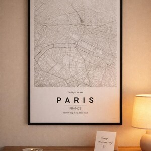May include: A monochrome map of Paris in a black frame, with the city name and coordinates. The text "The Night We Met" is above the city name. A small card with "Happy Anniversary" is on a wooden surface, next to a lamp.