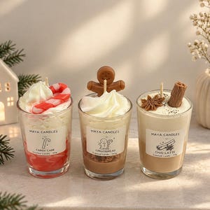 May include: Three glass jar candles by Maya Candles. The left candle is red and white with a candy cane topper. The centre candle is brown with a gingerbread man topper. The right candle is tan with star anise and cinnamon stick toppers.