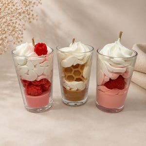 May include: Three glass candles designed to look like dessert parfaits. The left candle has pink wax, red raspberries, and white whipped wax. The middle candle has tan wax, a honeycomb shape, and white whipped wax. The right candle has pink wax, a strawberry, and white whipped wax.