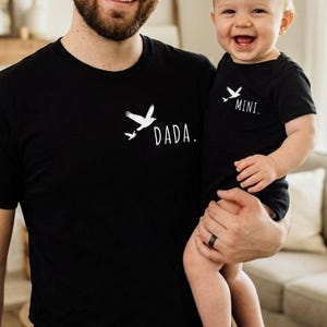 Peut inclure: Un t-shirt noir à manches courtes avec un motif blanc représentant deux oiseaux et le texte DADA. et un body pour bébé noir assorti à manches courtes avec deux oiseaux blancs et le texte MINI. Les deux vêtements ont un col rond.