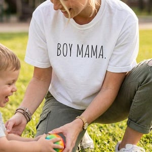 Peut inclure: Un t-shirt blanc à manches courtes avec le texte noir BOY MAMA. imprimé dans une police sans empattement simple sur la poitrine. Le vêtement présente une encolure ronde et une coupe standard, fabriqué dans un tissu blanc uni avec lettrage noir.