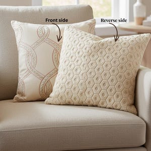 May include: Two decorative throw pillows on a beige sofa. One pillow features a cream-colored chain-link design with pink accents. The other pillow has a geometric honeycomb pattern in a cream color. The pillows add a touch of elegance to the living room.