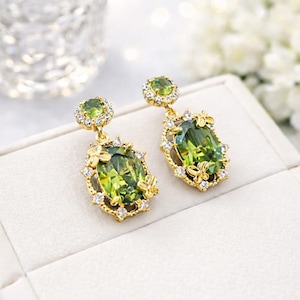 May include: A pair of gold-coloured drop earrings featuring oval, light green gemstones. The earrings have a halo of small, clear crystals and delicate floral accents. The earrings are displayed on a white surface.