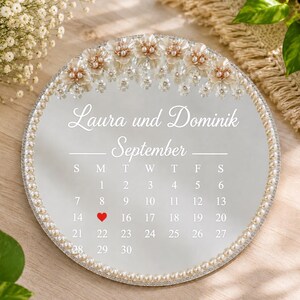 Op de afbeelding: Een ronde spiegelkalender voor september met de namen Laura und Dominik in witte letters. De bovenrand is versierd met witte bloemdecoraties en parels. Een rood hartje markeert de 15e dag en de rand is afgezet met parels.