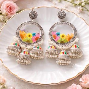 May include: Silver-toned earrings with a clear circular design filled with colorful dried flowers. Each earring features a dangling cluster of small silver bells with colorful accents and small white beads. The earrings are displayed on a white plate.