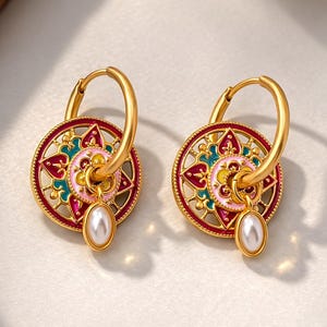 May include: A pair of gold-colored hoop earrings. Each earring features a circular, ornate design with red, pink, yellow, and teal accents. A white, oval-shaped pearl dangles from the center of each earring. The earrings are displayed on a light-colored surface.