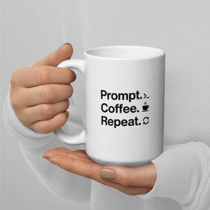 May include: White ceramic mug with the text "Prompt. > Coffee. ☕ Repeat. ♻" in black. The mug has a large handle and is held by a person.