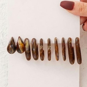 May include: A set of eleven brown and tan marbled resin earrings attached to a white display card. The collection includes two teardrop-shaped earrings and nine hoop earrings of varying sizes, all featuring a smooth, glossy finish.