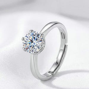 May include: A silver-toned adjustable ring featuring a single round-cut clear stone held in a six-prong setting. The band has a smooth, polished finish and includes a visible opening at the back to allow for size adjustment on the finger.