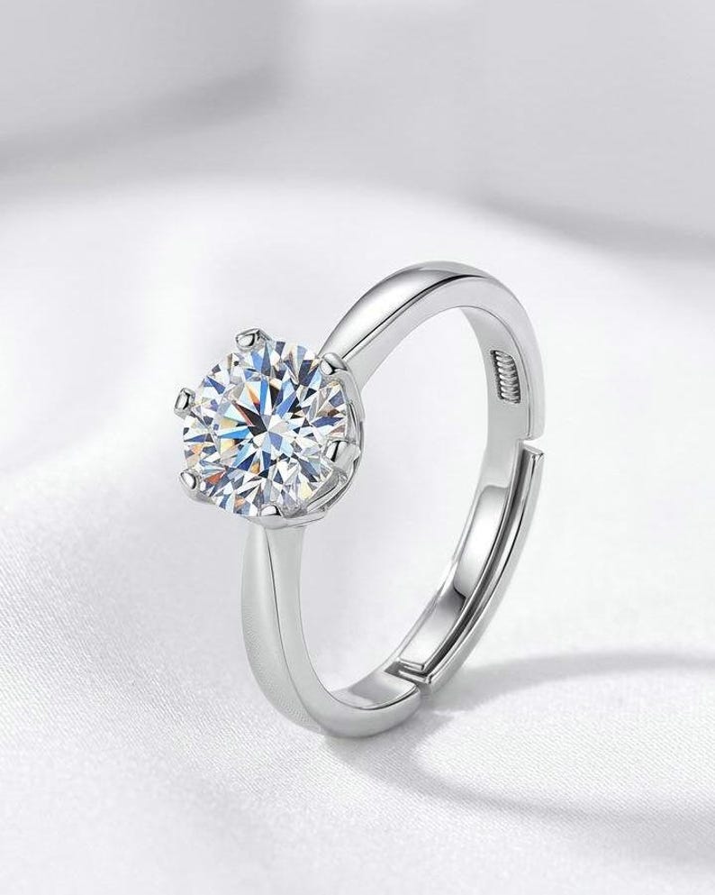 May include: A silver-toned adjustable ring featuring a single round-cut clear stone held in a six-prong setting. The band has a smooth, polished finish and includes a visible opening at the back to allow for size adjustment on the finger.