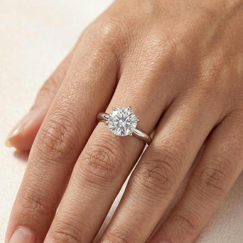 May include: A silver-toned ring featuring a single large, round, brilliant-cut clear gemstone set in a six-prong setting. The band is smooth and polished, showcasing a classic solitaire design with a high-shine metallic finish and clear stone.