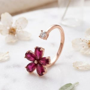 Könnte beinhalten: Roségoldener Blumenring mit tiefrosa Blütenblättern und einem klaren Edelstein. Das offene Design hat einen zweiten klaren Edelstein oben, was einen einzigartigen und eleganten Look ergibt.