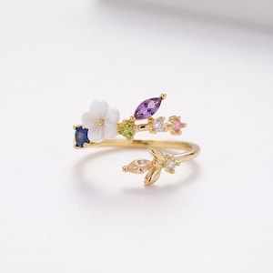 May include: A gold ring with a floral design, featuring a white flower and various coloured gemstones, including blue, purple, green, and pink. The ring's design is inspired by nature, with a band that wraps around the finger.