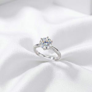 May include: A silver-toned ring featuring a single round-cut clear gemstone held in a six-prong setting. The band is smooth and polished, showcasing a reflective metallic finish that complements the brilliant, multi-faceted center stone.