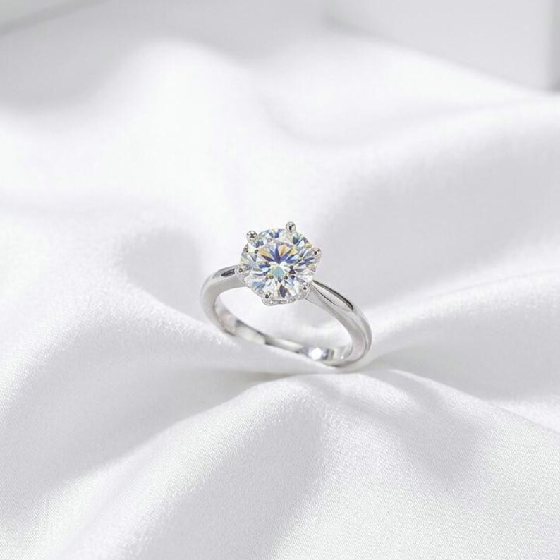 May include: A silver-toned ring featuring a single round-cut clear gemstone held in a six-prong setting. The band is smooth and polished, showcasing a reflective metallic finish that complements the brilliant, multi-faceted center stone.