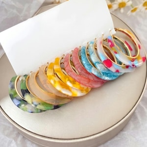 May include: A collection of hoop earrings attached to a white display card. Each hoop features a gold-tone metal frame and a colorful, marbled resin insert. The set includes various colors such as green, beige, yellow, pink, blue, and multi-colored patterns.