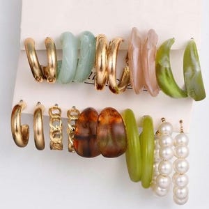 May include: A collection of twelve pairs of earrings displayed on a white card. The set includes gold-toned metal hoops, chain-link hoops, translucent resin hoops in shades of green, brown, and pink, and two pairs of white faux pearl drop earrings.