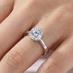 May include: A silver-toned ring featuring a single round, brilliant-cut clear stone held in a six-prong setting. The band is smooth, polished, and solid, showcasing a classic design with a high-shine metallic finish and a faceted center stone.